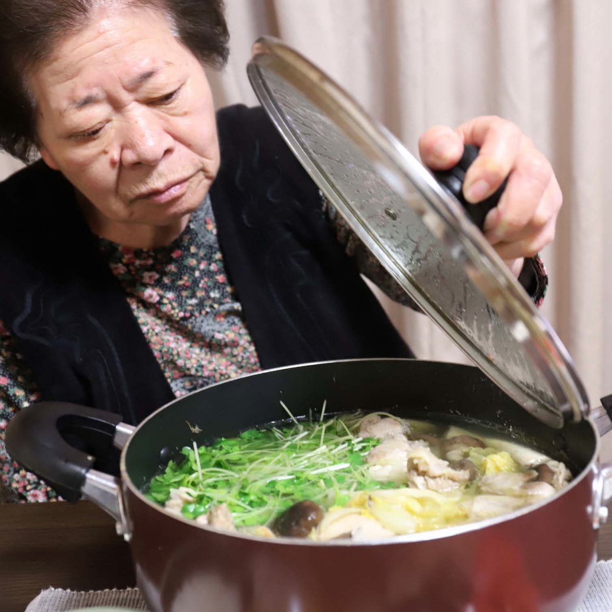食卓の水炊きの鍋のフタを片手で持ち上げている祖母（おばあ）