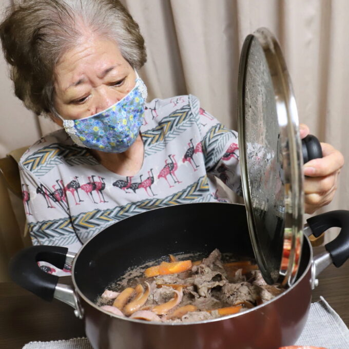 食卓に着いて、鍋のフタを開けている祖母・おばあ