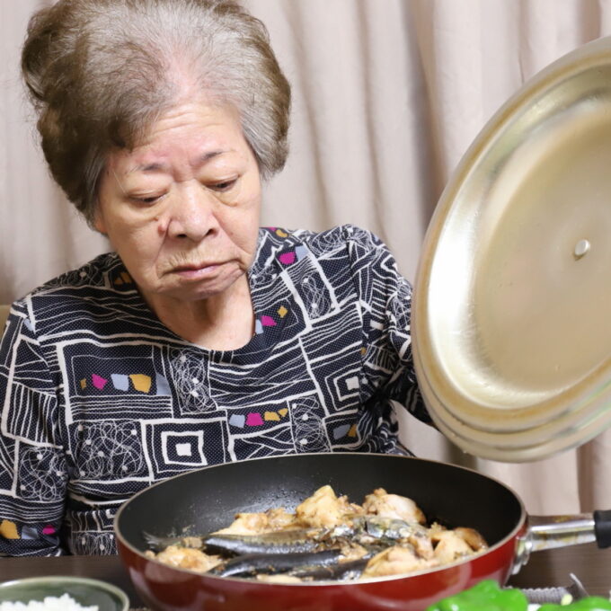 食卓でフライパンのフタを開けている祖母・おばあ