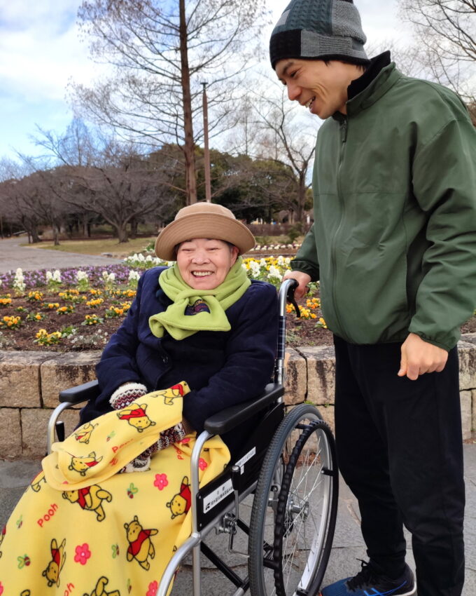 公園の花壇の前で車イスに乗って笑顔の祖母と付き添う孫