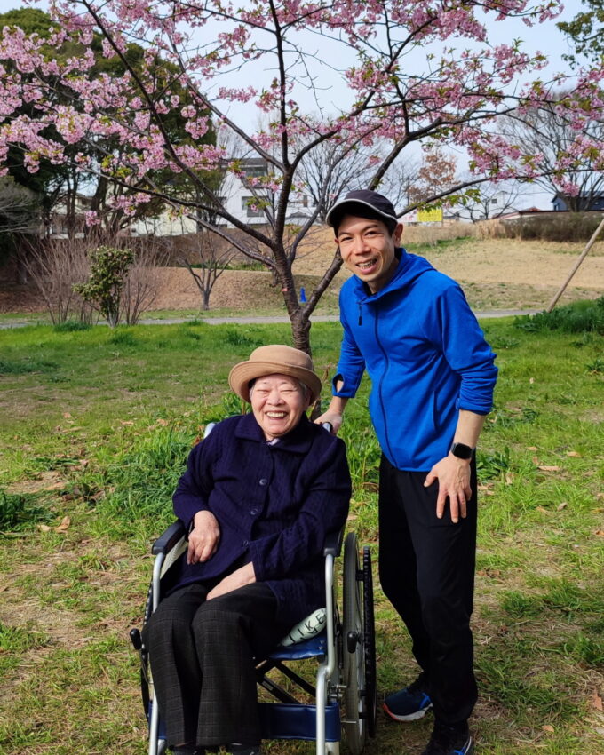 花が満開の桜の木の前にいる車イスに座った祖母と横に立つ孫