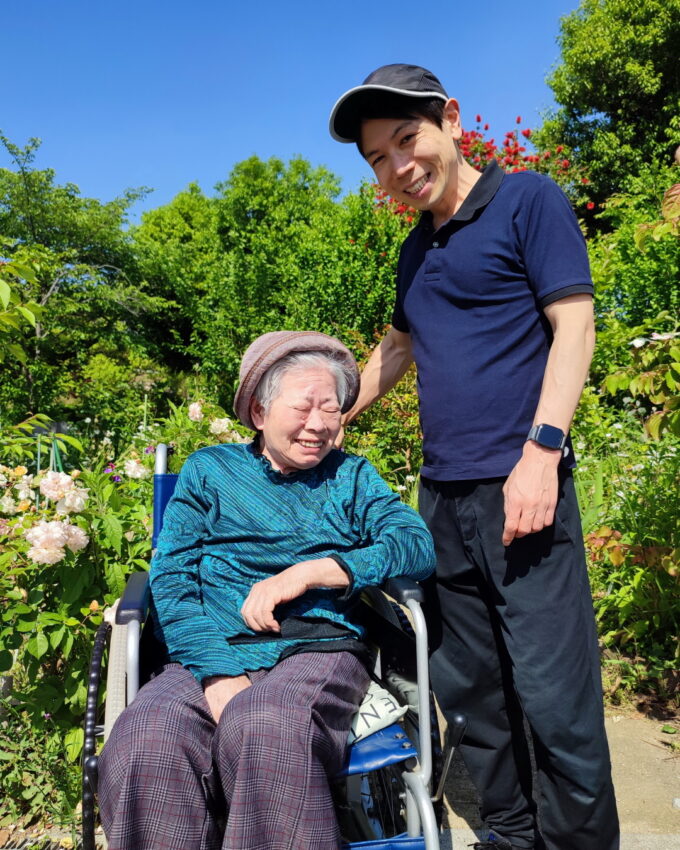 青空の下、花が咲く公園で車イスに乗って笑う祖母と、付き添う孫