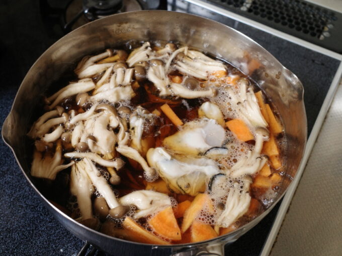 ステンレスの片手鍋に炊き込みご飯の具材、ニンジンやシメジや牡蠣を入れて煮ている
