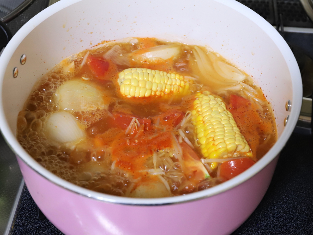 鍋に玉ねぎやニンジン、トマト、トウモロコシなどを入れて煮てキムチ鍋を作っている