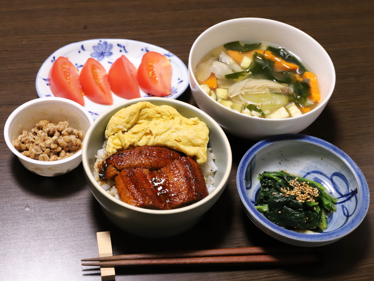 ごはんにうなぎと卵を乗せたウナ玉丼、ホウレンソウのおひたし、味噌汁、納豆、トマトというお皿に並べた晩ごはんの献立